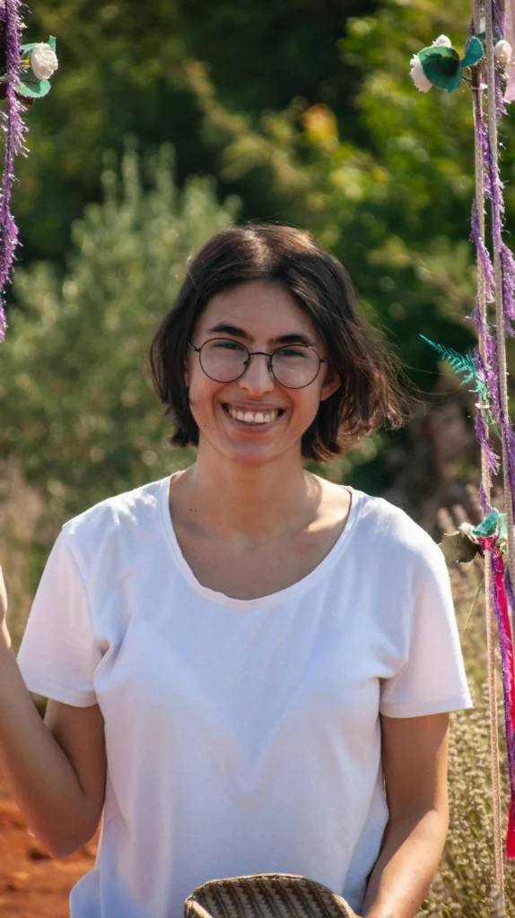 Mulher jovem sorrindo ao ar livre, usando óculos e camiseta branca, em ambiente natural decorado com fitas coloridas.