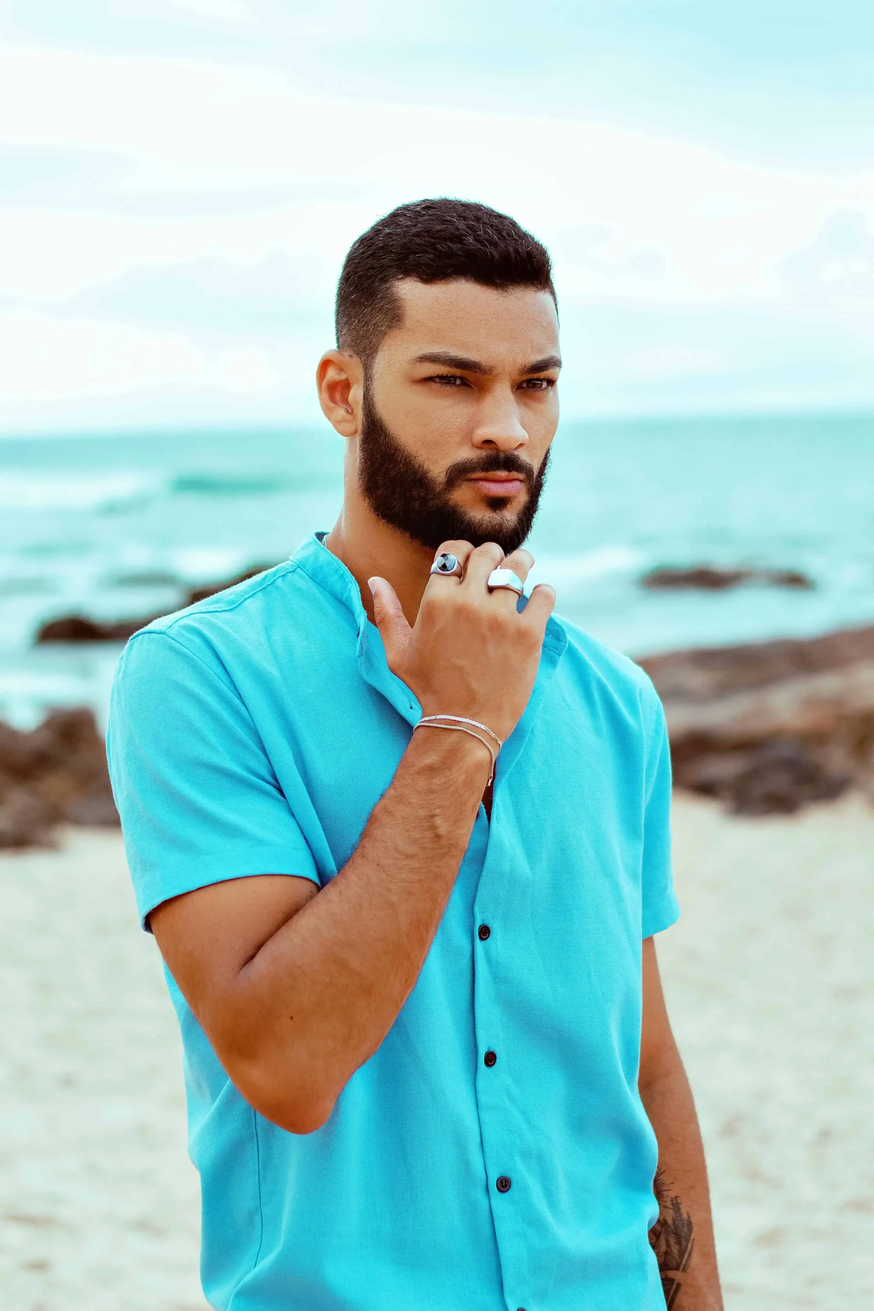 Homem de barba usando camisa azul clara em uma praia, olhando seriamente para o horizonte com o mar ao fundo.