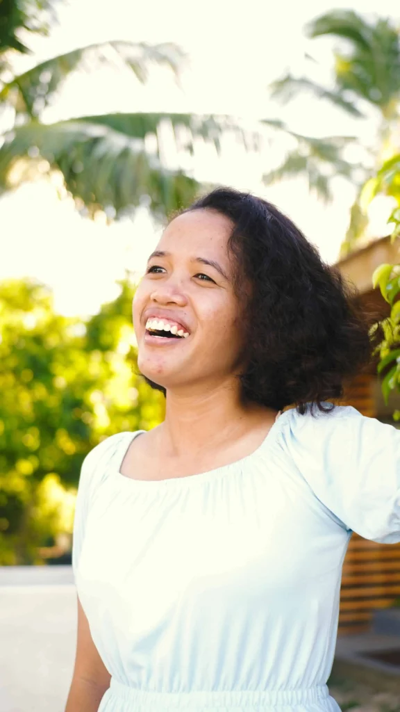 Mulher sorrindo ao ar livre em ambiente tropical, usando vestido claro, com árvores e palmeiras ao fundo.