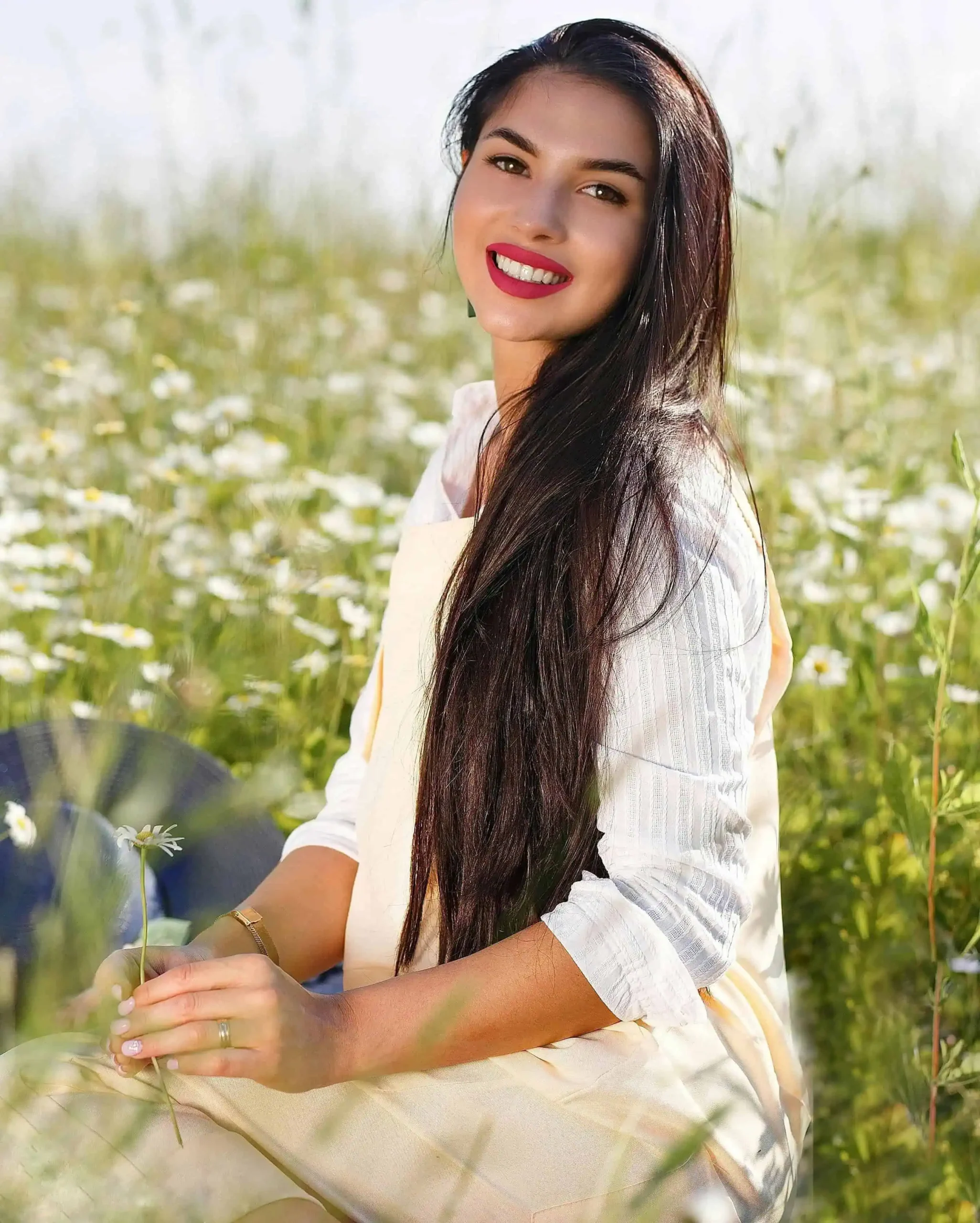 Mulher jovem de cabelos longos e escuros sorrindo em um campo florido enquanto segura uma pequena flor branca.