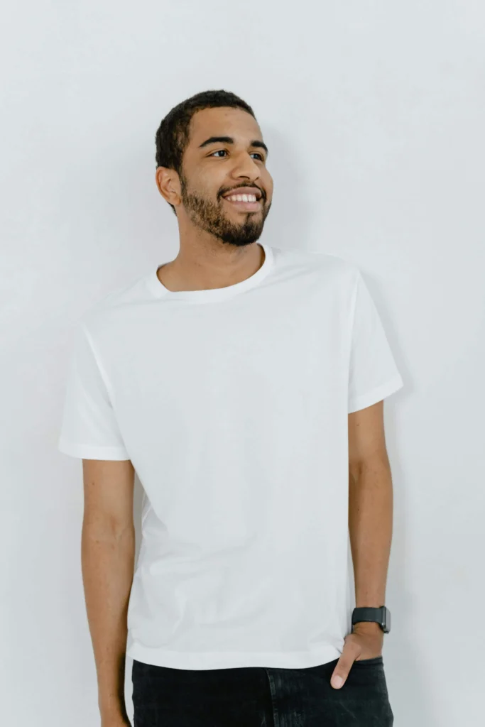 Homem jovem sorrindo, usando camiseta branca, em pé diante de um fundo claro e minimalista.