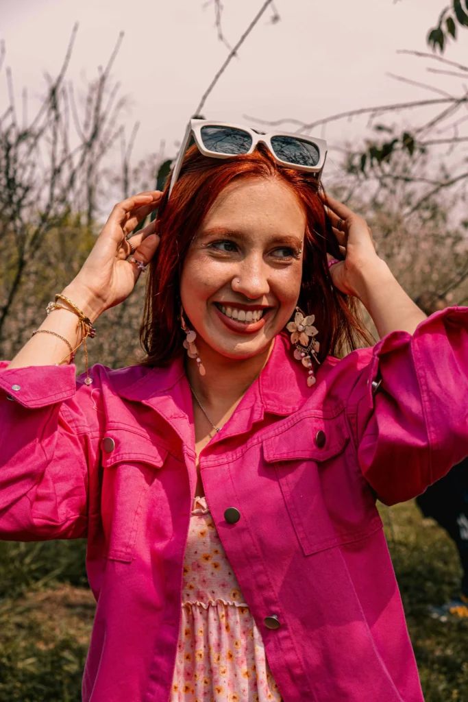 Kabbalista Miriam sorrindo ao ar livre, usando jaqueta rosa vibrante e óculos sobre a cabeça, representante espiritual do mês hebraico de Adar.