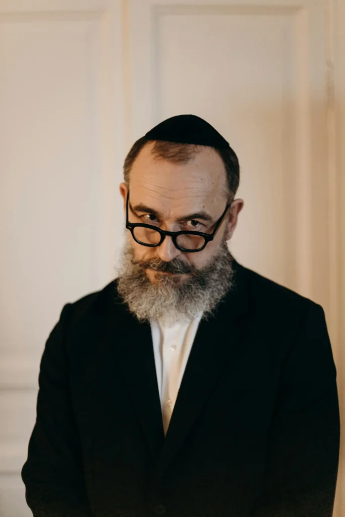 Kabbalista Yosef com barba grisalha, óculos e vestes escuras tradicionais, representante espiritual do mês hebraico de Adar.